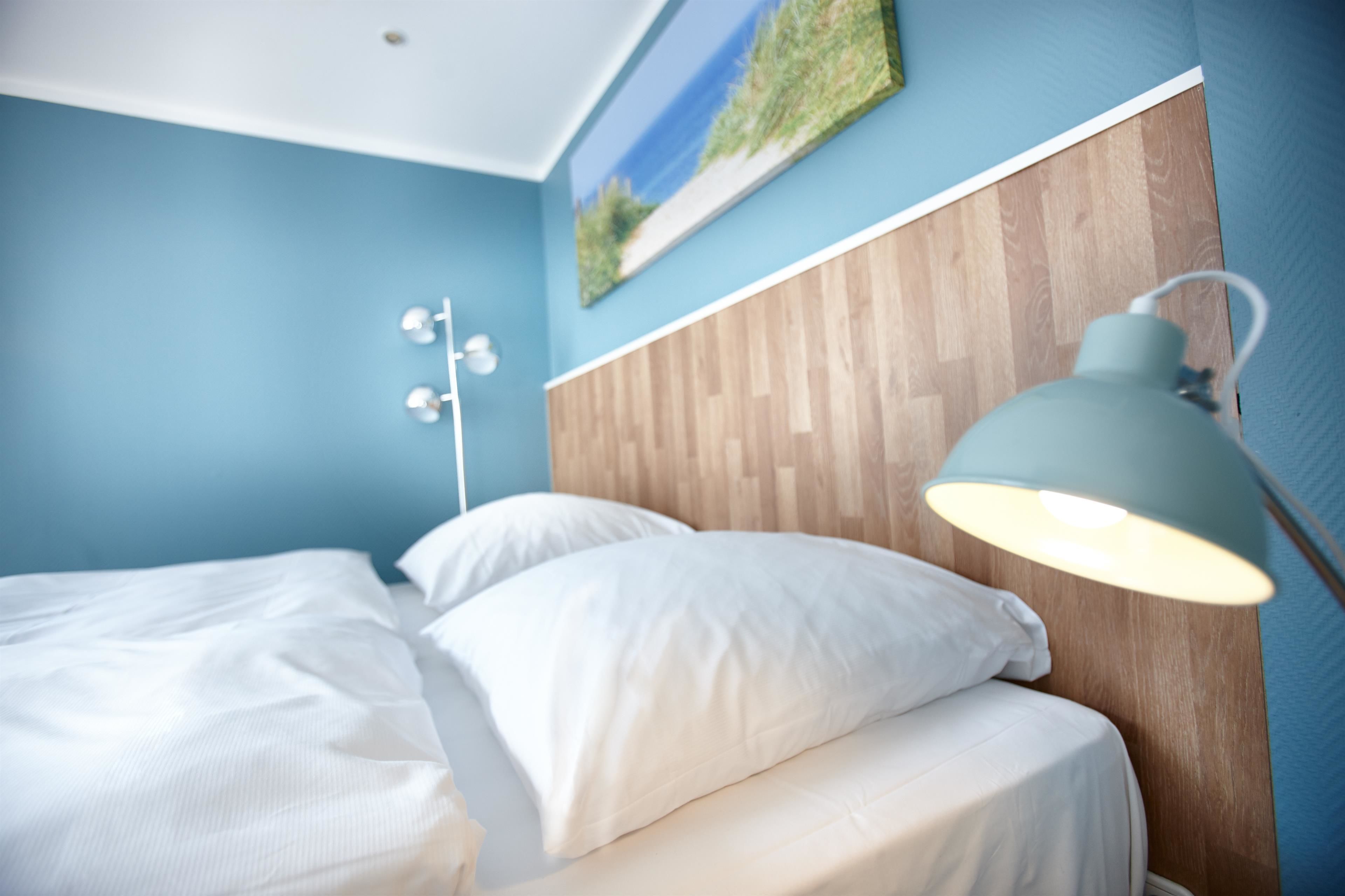 A bedroom with a bed, white pillows, a wooden headboard, a wall-mounted lamp, and a painting of a landscape.