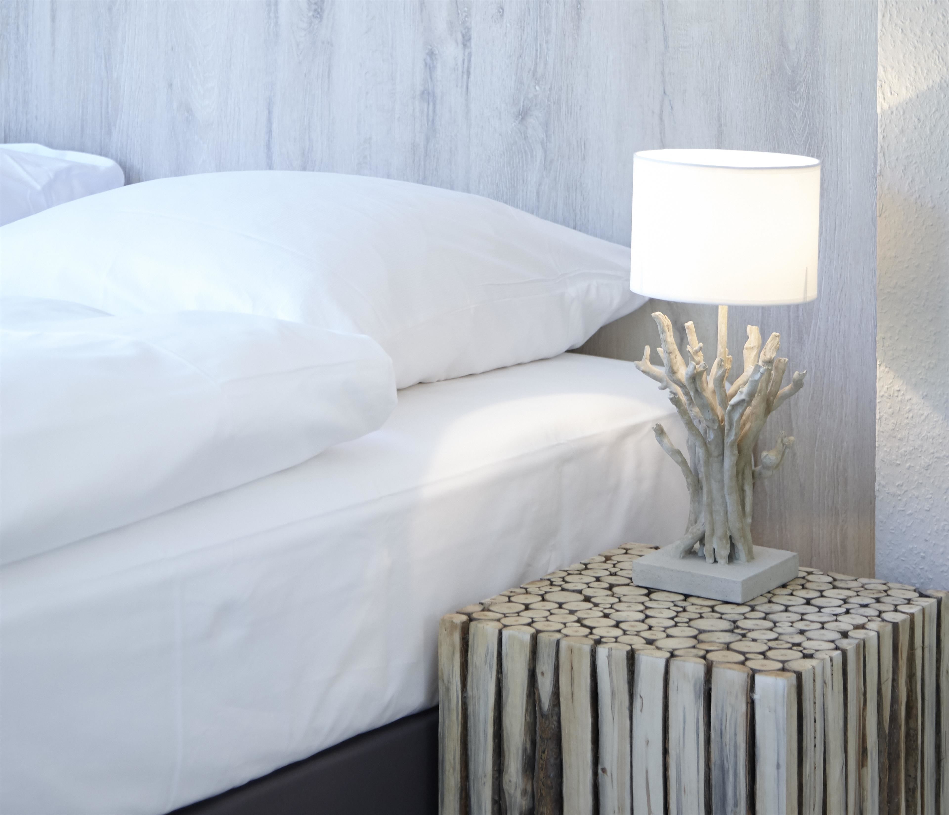 Una cama bien hecha con ropa de cama blanca, una lámpara blanca sobre una mesita de noche de madera y una pared con textura.