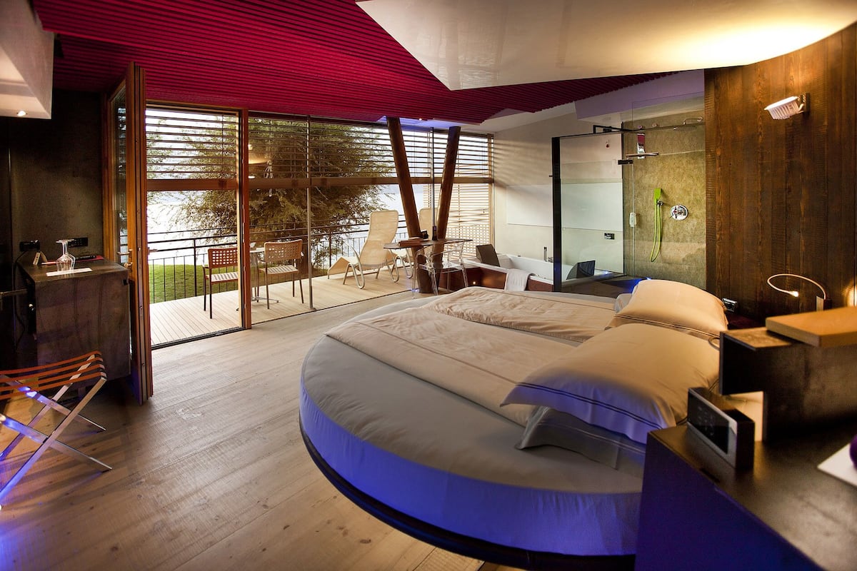 A modern bedroom with a curved bed, a glass shower, and a balcony with a view of trees.