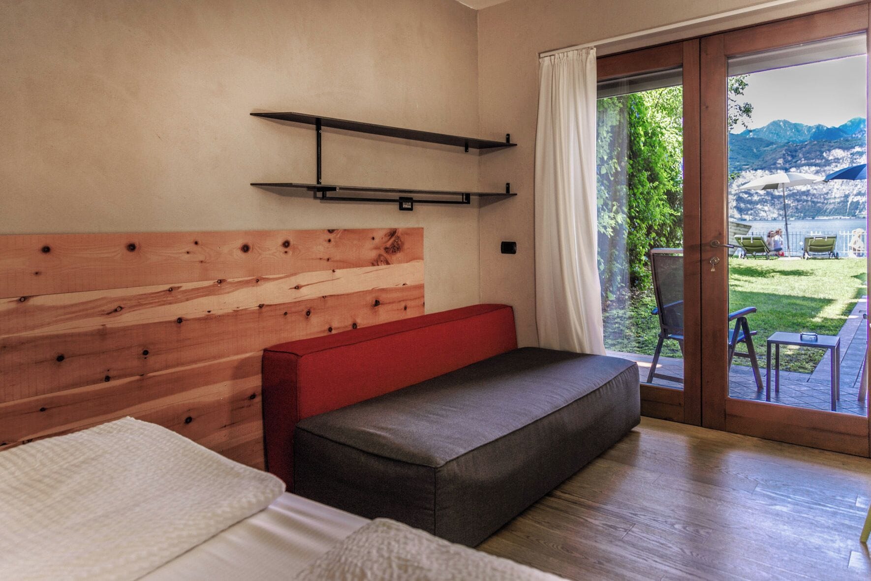 Une chambre d’hôtel avec une tête de lit en bois, un banc rouge, un lit et une vue sur un jardin et des montagnes, visible à travers une porte vitrée.