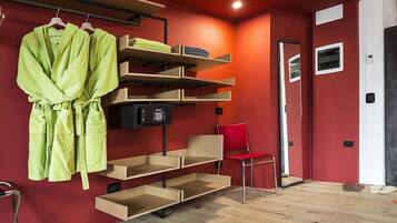 Habitación de hotel con pared roja, suelo de madera, una silla roja, un espejo y un estante con bata y toallas.