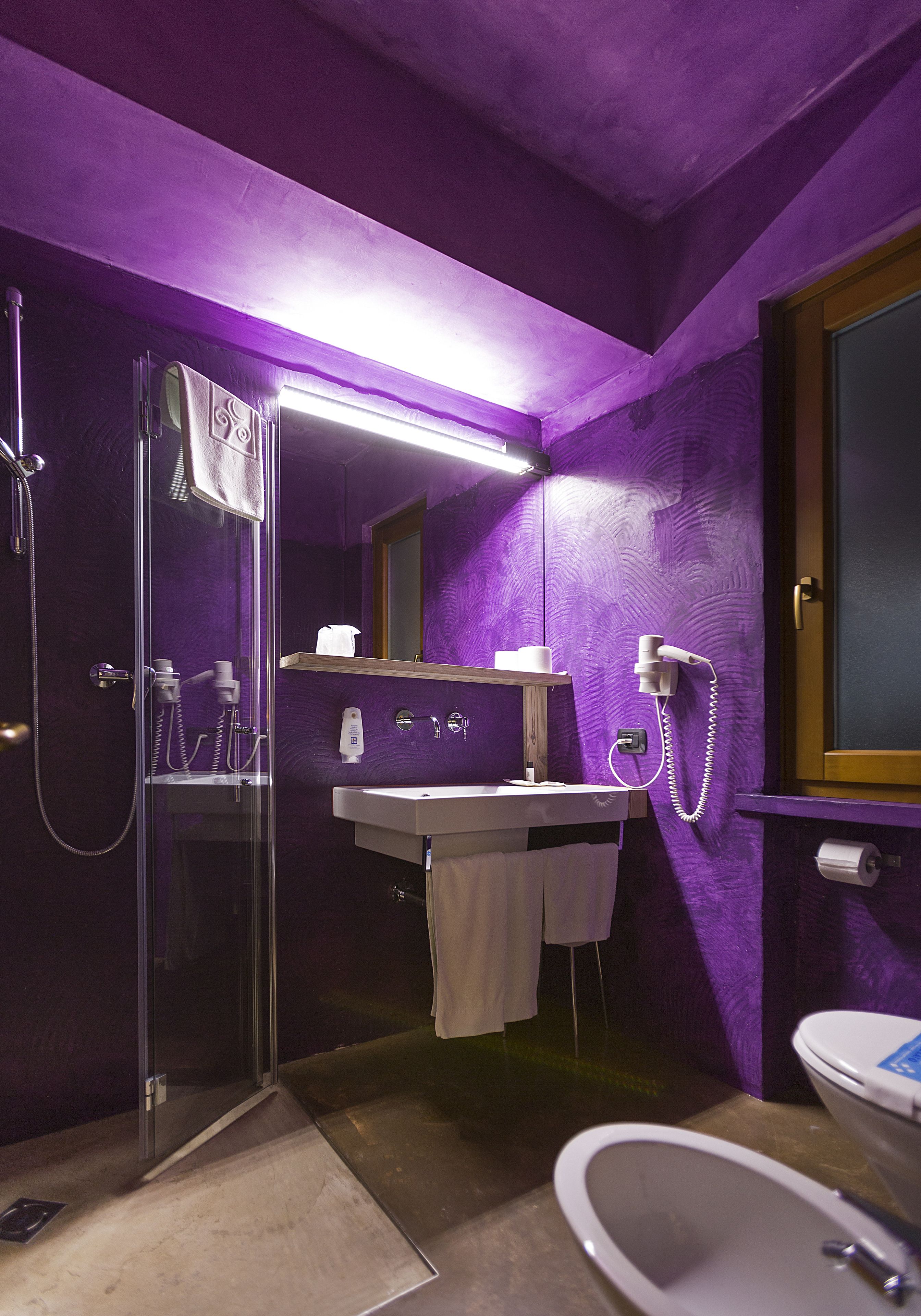 Une salle de bain aux murs violets, dotée d’une douche en verre, d’un lavabo avec un miroir, de toilettes et d’un téléphone fixé au mur.