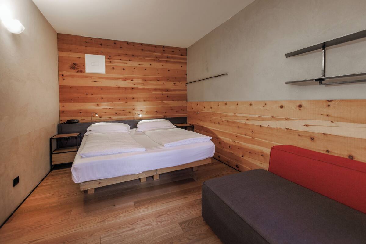 A room with a wooden wall, a bed with white bedding, a red bench, and a small table with a lamp.