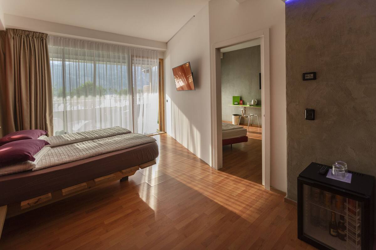 A modern hotel room with a wooden floor, a bed with pillows, a TV mounted on the wall, and a small table with a glass.
