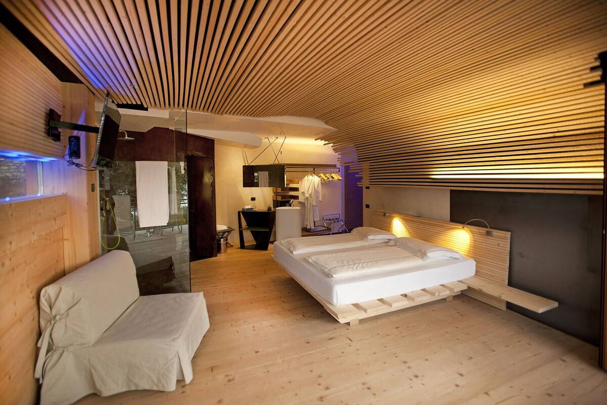 A modern hotel room with a wooden ceiling, a large bed, a seating area, and a bathroom visible through a glass partition.