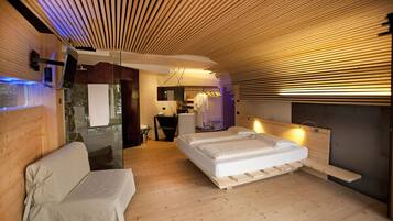 A modern hotel room with a wooden ceiling, a large bed, a seating area, and a bathroom visible through a glass partition.