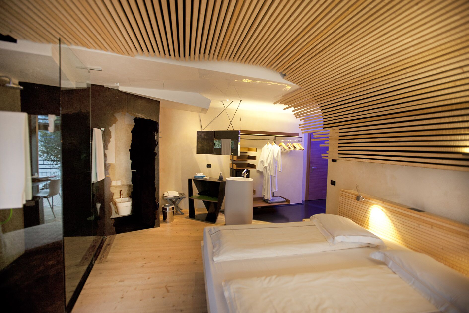 A modern bedroom with a wooden slatted ceiling, a large bed, a vanity with a mirror, and a hanging robe rack.