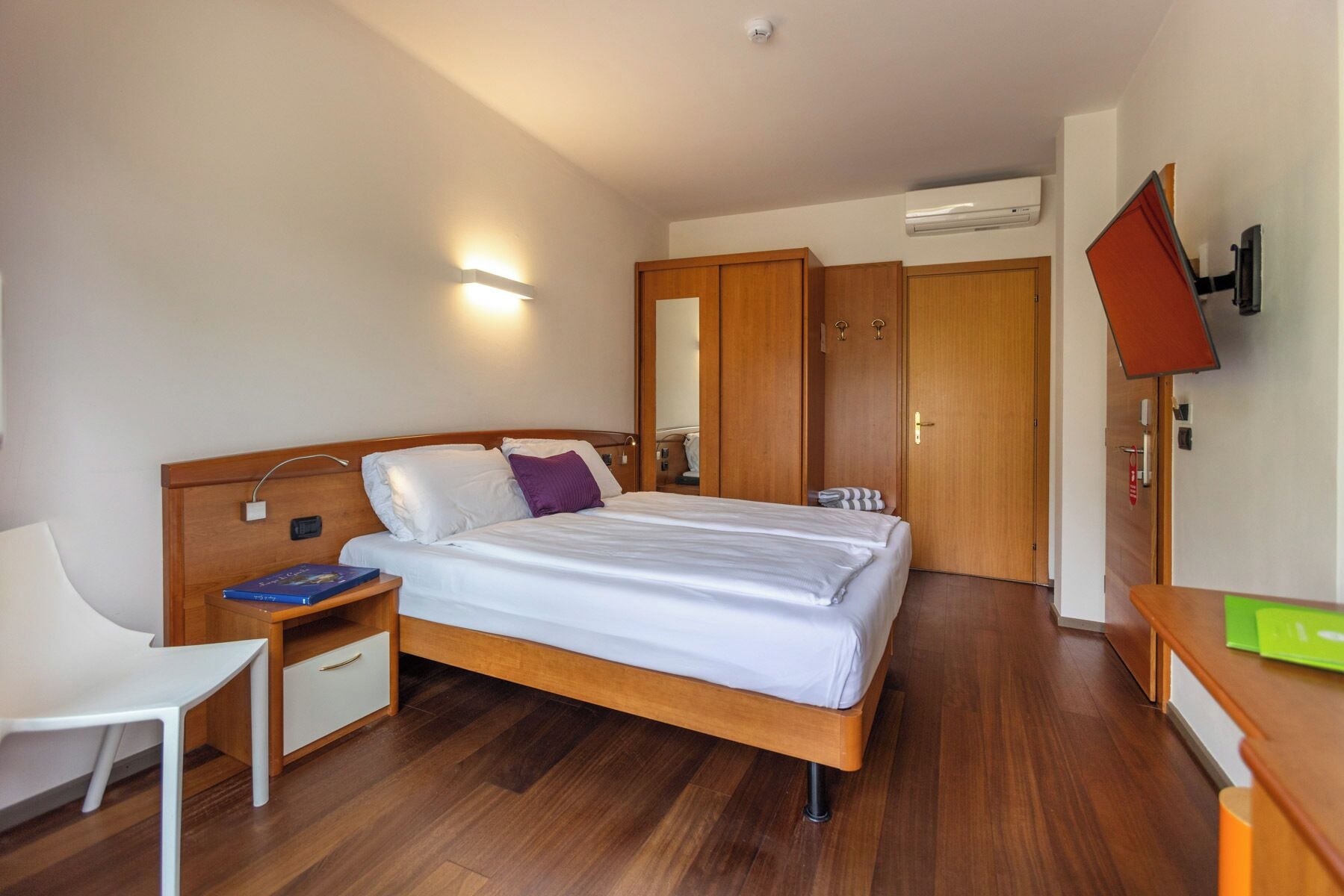 A hotel room with a bed, bedside table, chair, wardrobe, and a TV.
