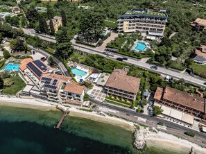 An aerial view of a coastal hotel with multiple buildings, swimming pools, and a pier extending into the water. - Ambienthotel Spiaggia (Malcesine)