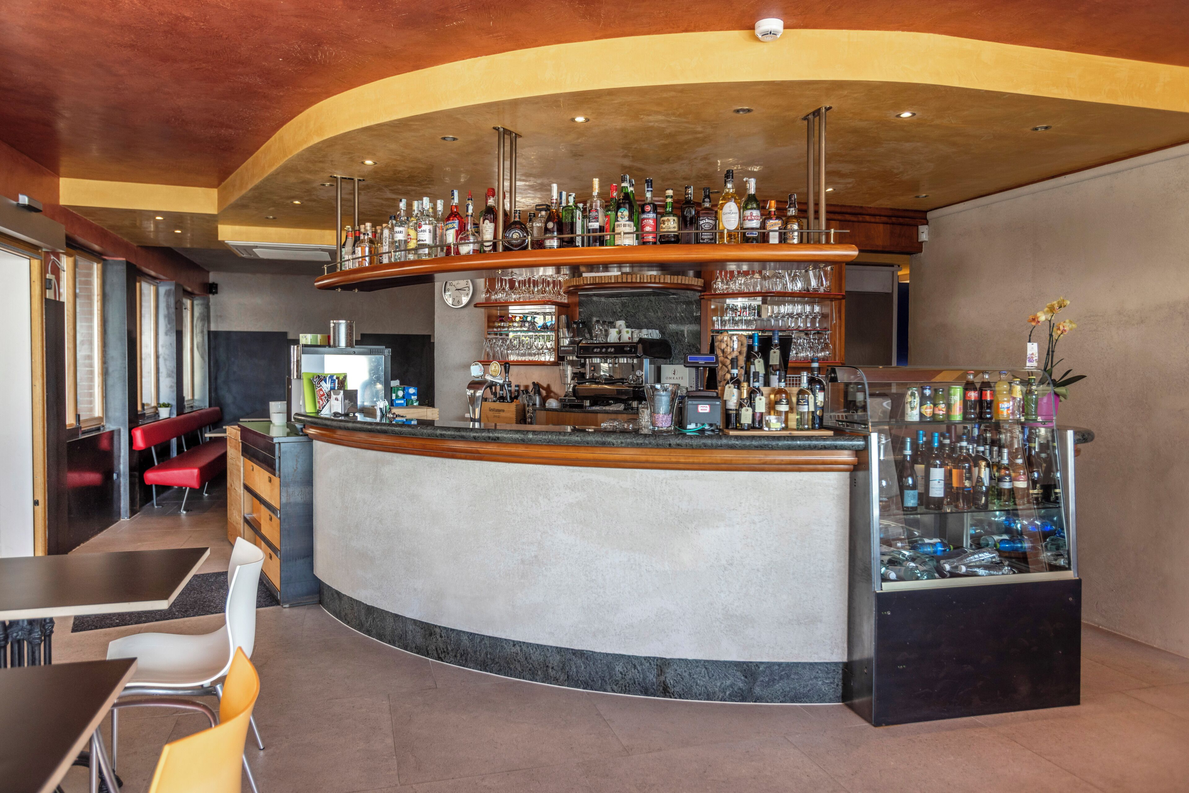 Un'area bar con un bancone curvo, scaffali pieni di bottiglie, bicchieri e una vetrina. Sono presenti zone con tavoli e sedie.