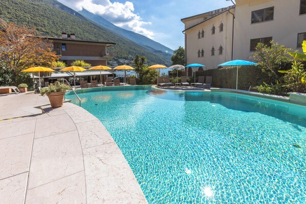 A swimming pool with lounge chairs and umbrellas, surrounded by buildings and mountains. - Ambienthotel Spiaggia (Malcesine)