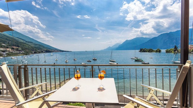 Una terraza con mesa y sillas, con vista a un lago con barcos, rodeada de montañas.