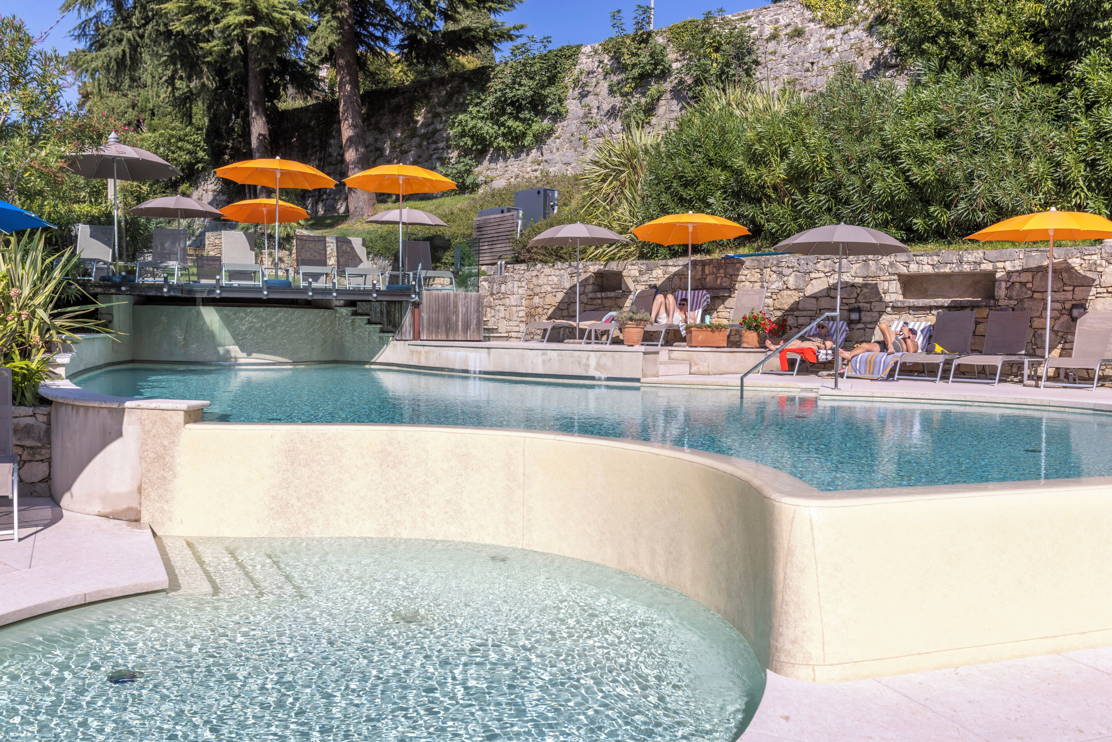 A pool with multiple umbrellas, lounge chairs, and a stone wall.