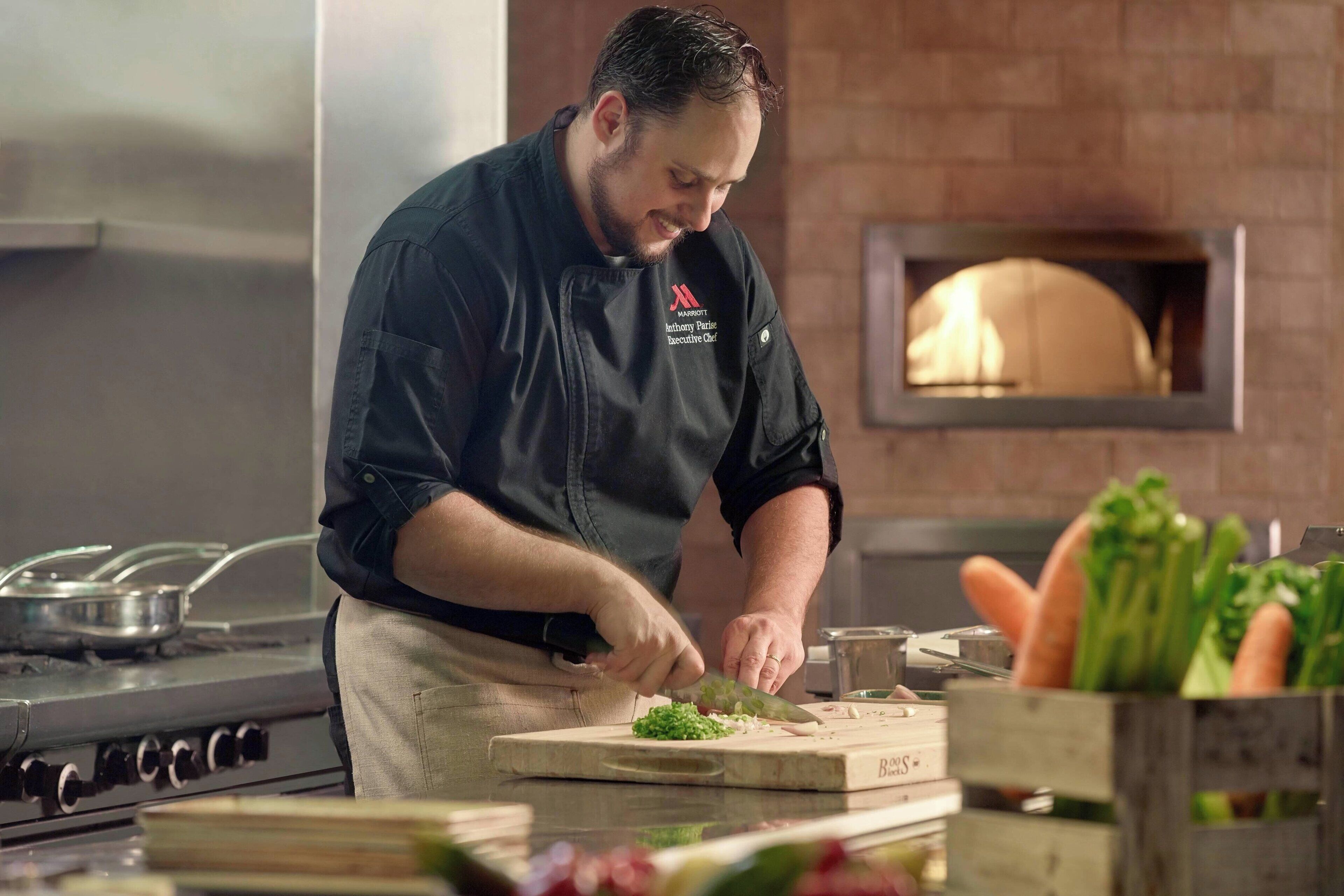 A chef in a professional kitchen with a brick wall, a stove, and a pizza oven.