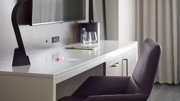 A modern hotel room with a white desk, a flat-screen TV, a water bottle, and a glass.