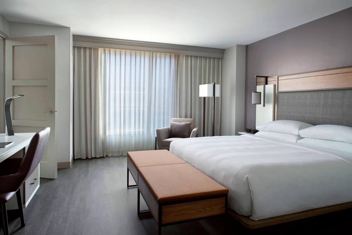 A hotel room with a large bed, a desk, a chair, a bench, and a window with curtains.