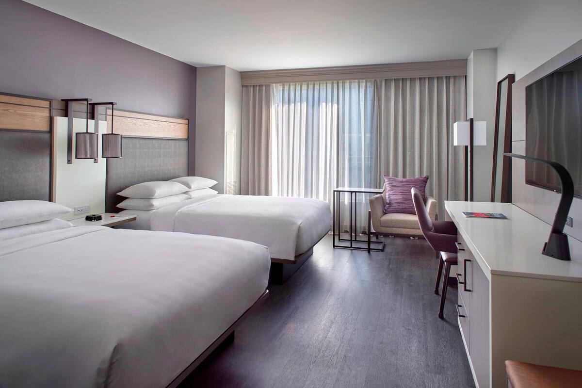 A hotel room with two beds, a desk, a chair, and a large window with curtains.