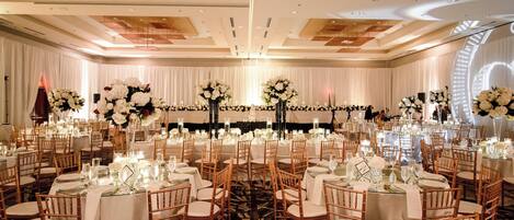 Un salón de banquetes con mesas redondas, manteles blancos y centros de mesa florales.