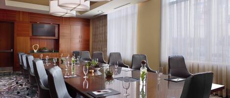 A conference room with a long table, multiple chairs, a flat-screen TV, and a large window with sheer curtains.