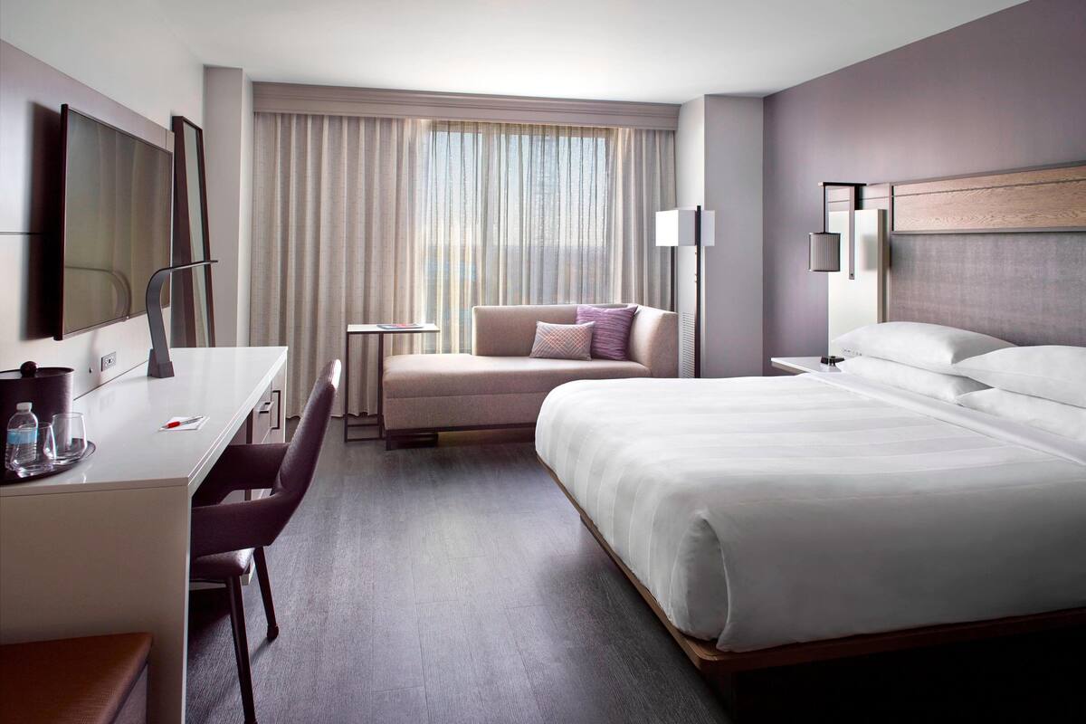 A hotel room with a bed, a desk, a chair, a small table, and a large window with curtains.