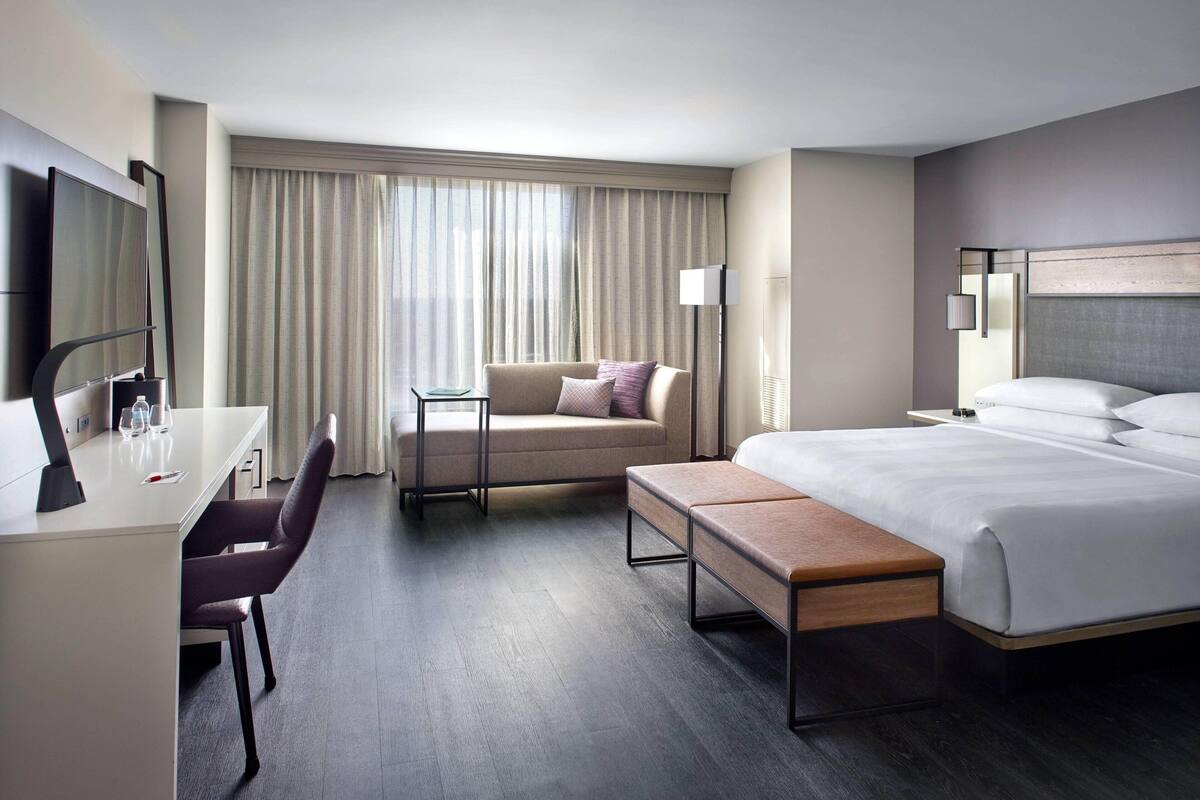 A modern hotel room with a bed, a desk, a chair, a sofa, and a large window with curtains.