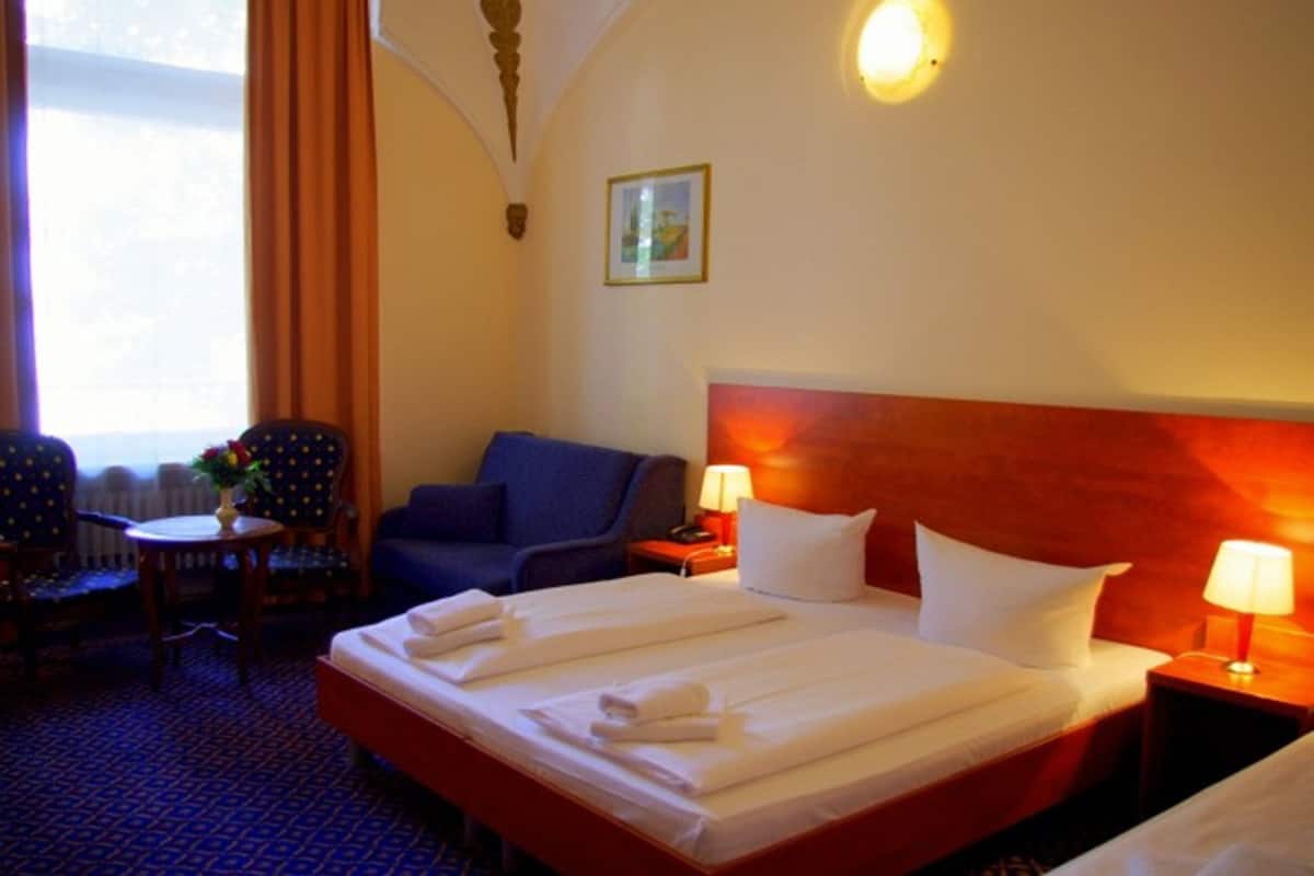 Une chambre d’hôtel avec un grand lit, un fauteuil bleu, une petite table avec un vase et un tableau au mur.