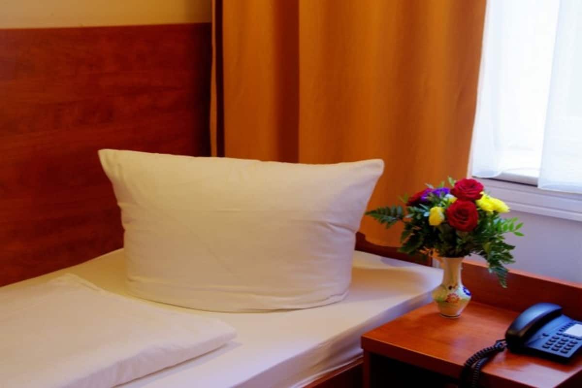 Une chambre d’hôtel avec un lit, un oreiller blanc, un vase de fleurs et un téléphone posé sur une table de chevet en bois.