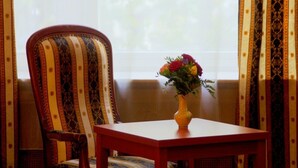 A chair with a striped pattern, a table with a vase of flowers, and curtains with a floral design. - City Hotel am Kurfürstendamm (Berlin)