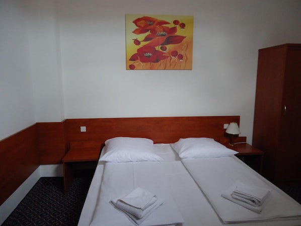 A hotel room with two single beds, a wooden headboard, a nightstand with a lamp, and a painting on the wall.