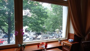 A dining area with a view of a busy street through large windows, a table with a vase of flowers, and a small potted plant on the windowsill. - City Hotel am Kurfürstendamm (Berlin)
