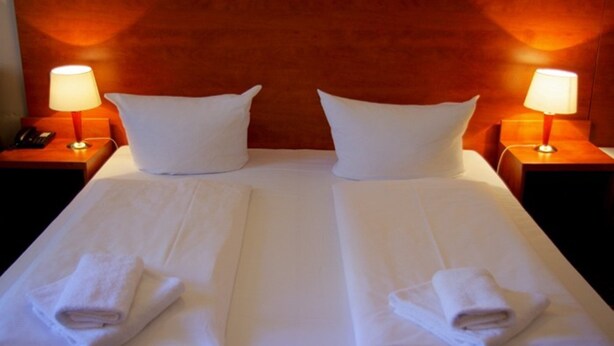 A double bed with white linens and two pillows, flanked by nightstands with lamps, in a room with wooden paneling. - City Hotel am Kurfürstendamm (Berlin)