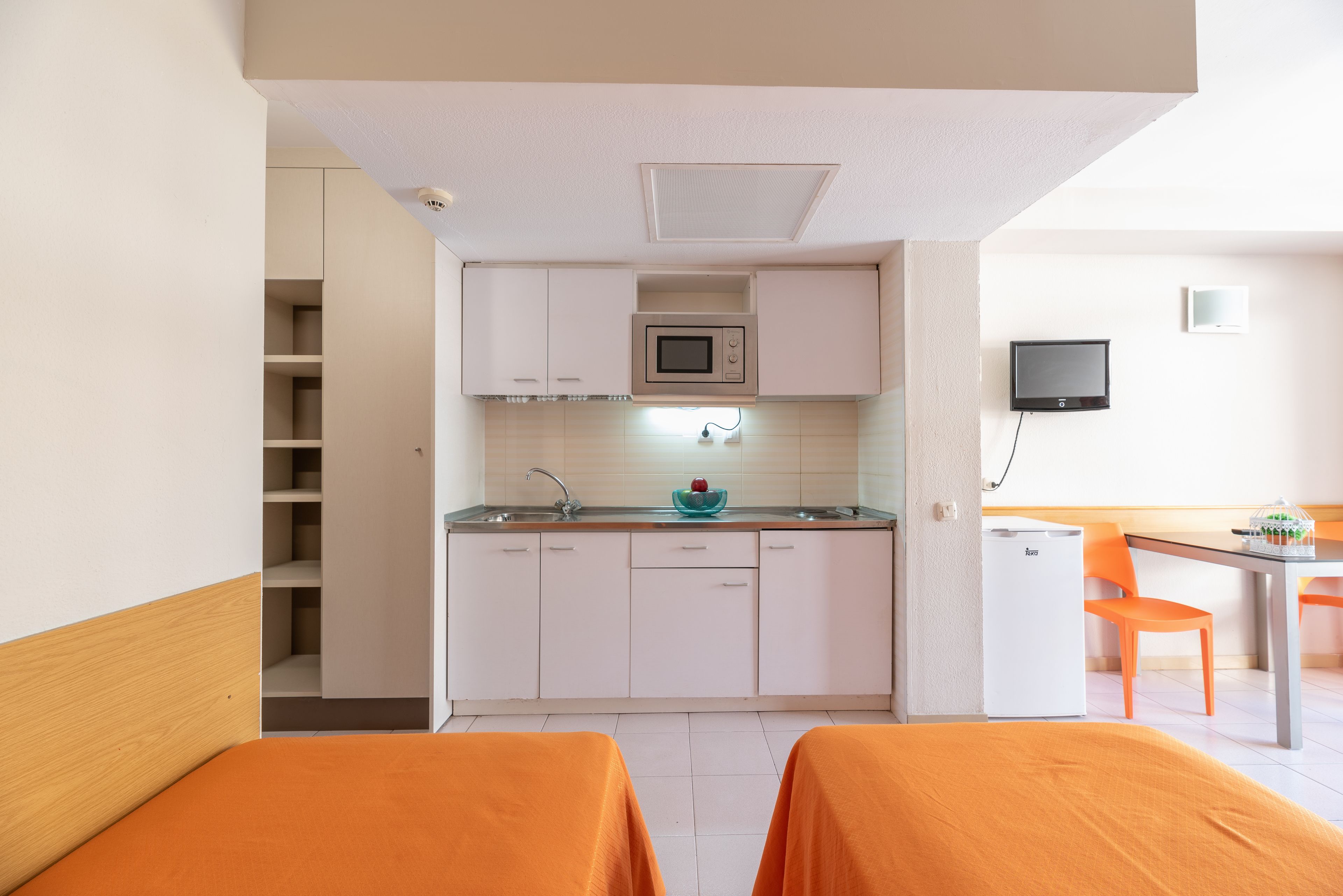 A compact hotel room with a kitchenette, a bed with orange bedding, a dining area with a table and chairs, and a television mounted on the wall.