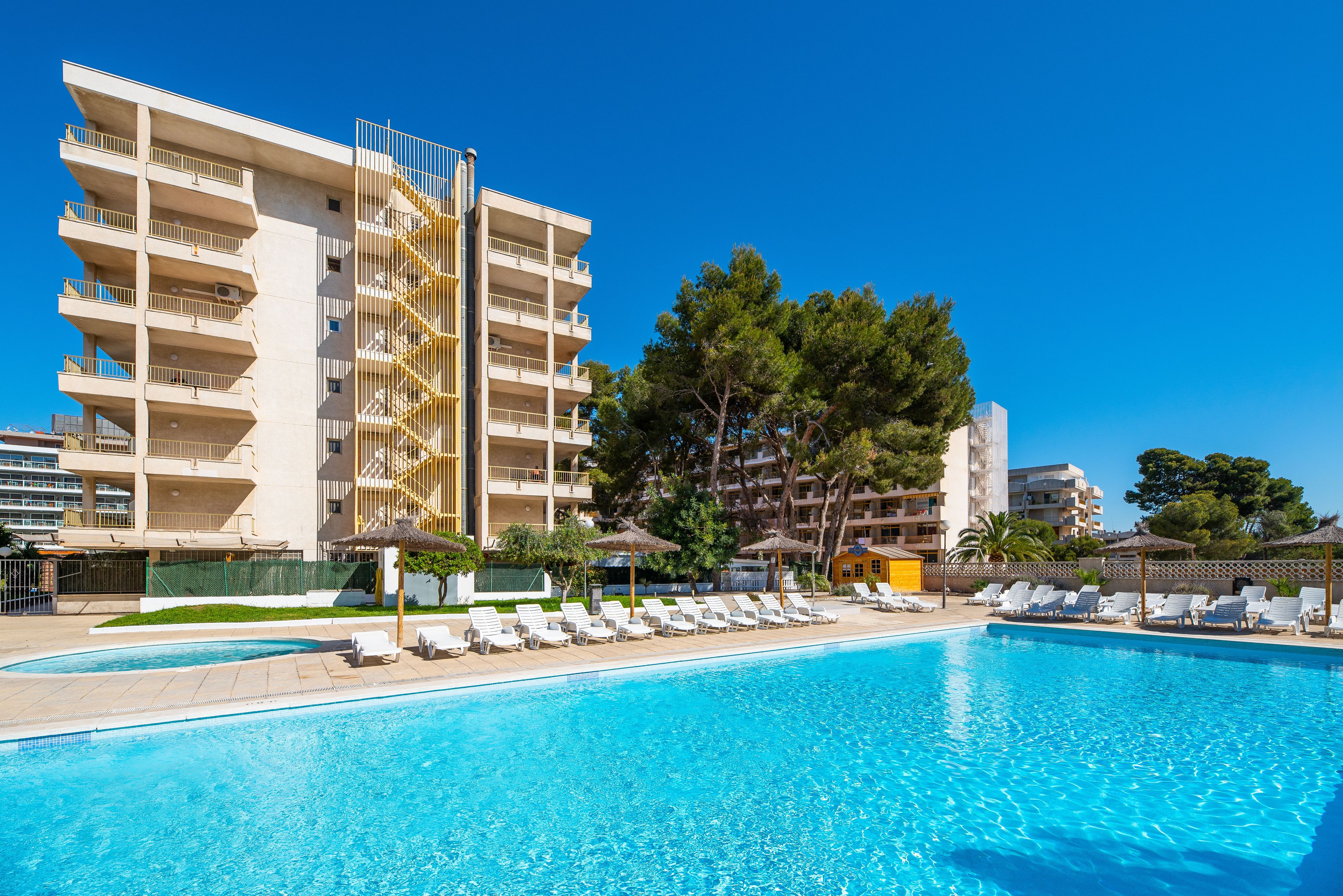 Une piscine entourée de chaises longues et de parasols, elle-même bordée d’un immeuble à plusieurs étages doté de balcons.
