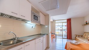 A modern kitchen with a built-in microwave, stainless steel sink, and white cabinets. There is a dining area with a table and chairs, and a bed with an orange bedspread. - Rentalmar Salou Pacific (Salou)