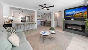 A modern living room with a sofa, a coffee table, and a TV mounted on the wall. There is a kitchen in the background with a dining area.