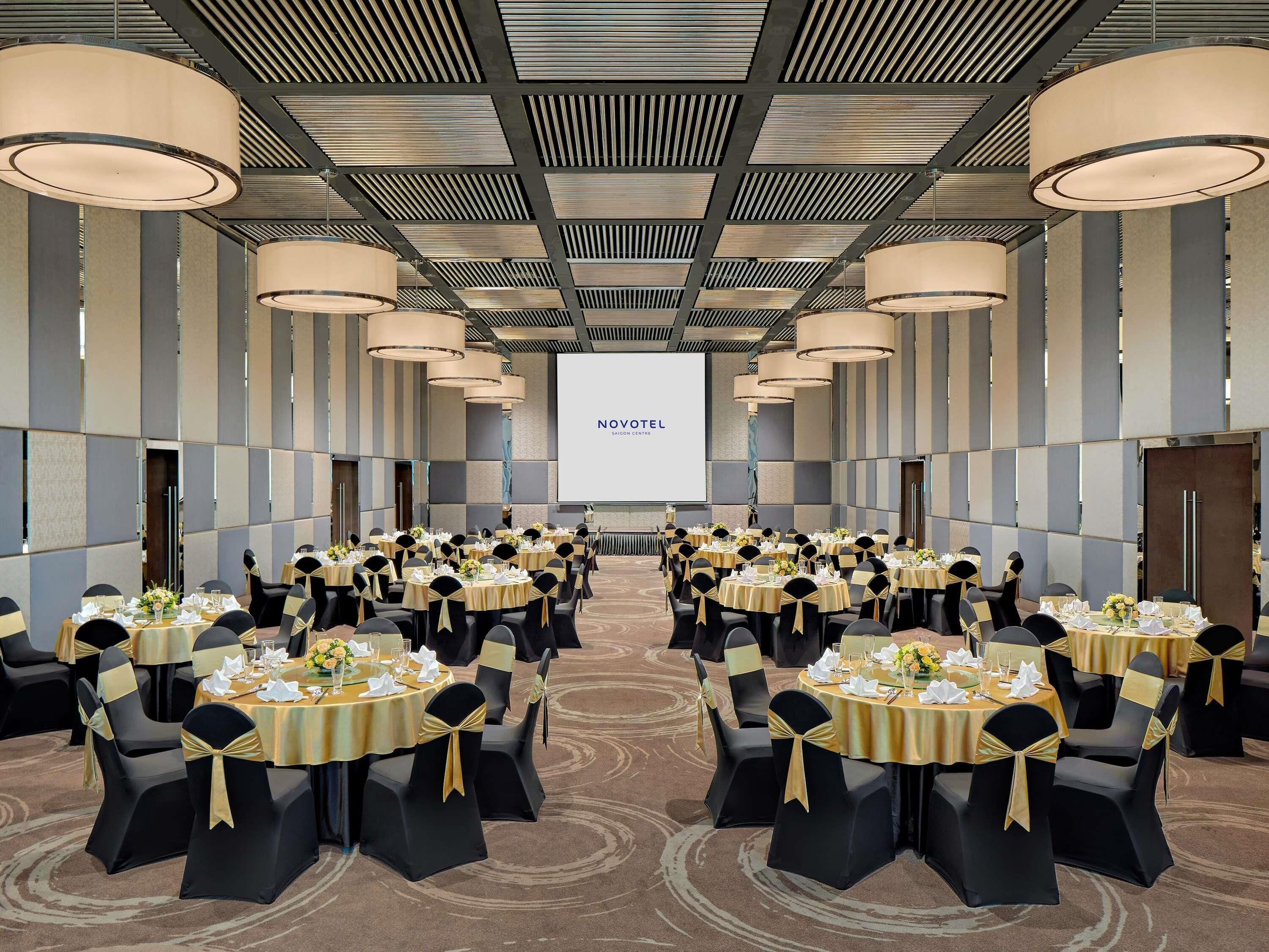 A conference room with round tables, black chairs with gold sashes, and a large screen displaying 'NOVOMATIC SAIGON CENTER'.