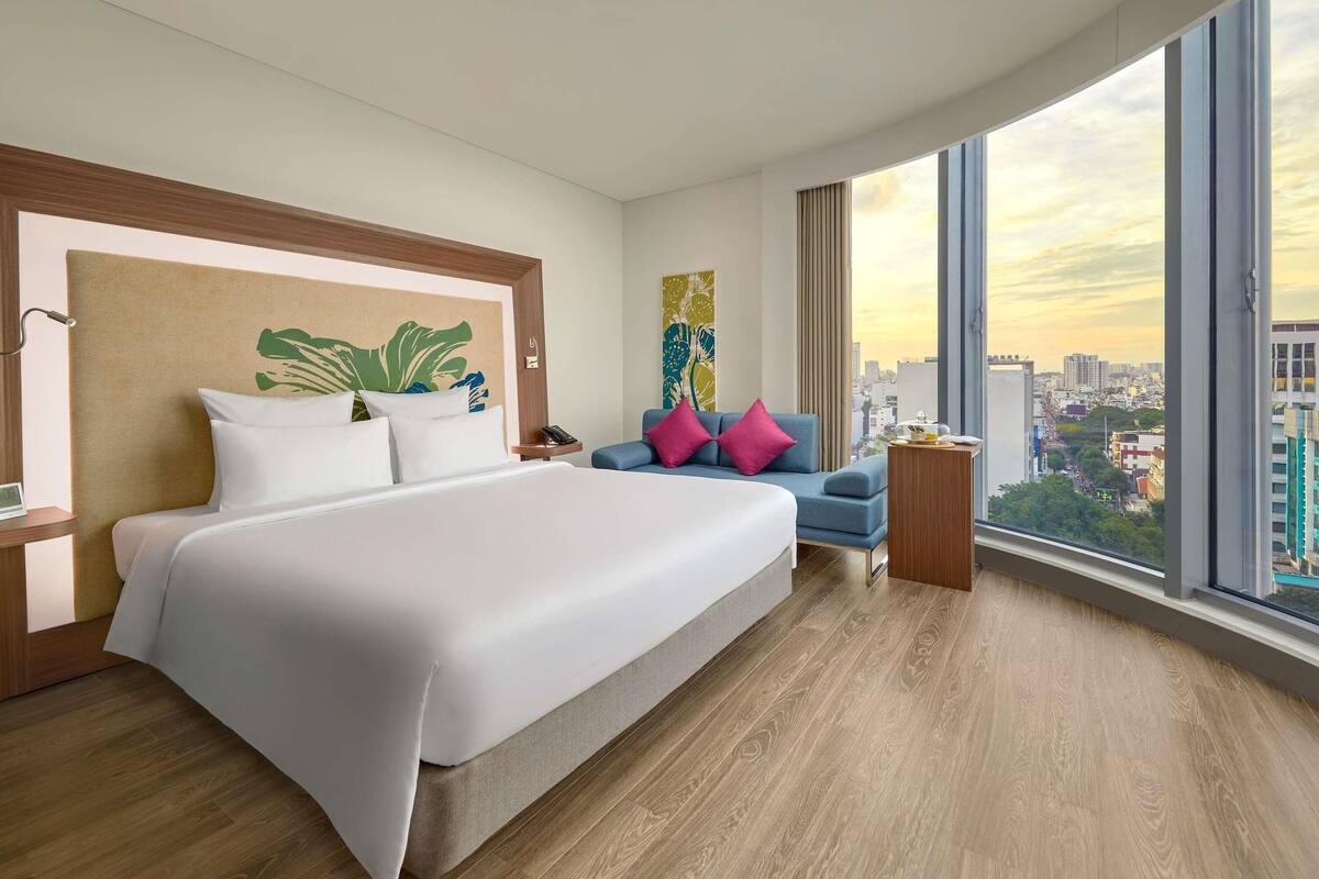 Een moderne hotelkamer met een groot bed, een blauwe fauteuil en uitzicht op de stad.