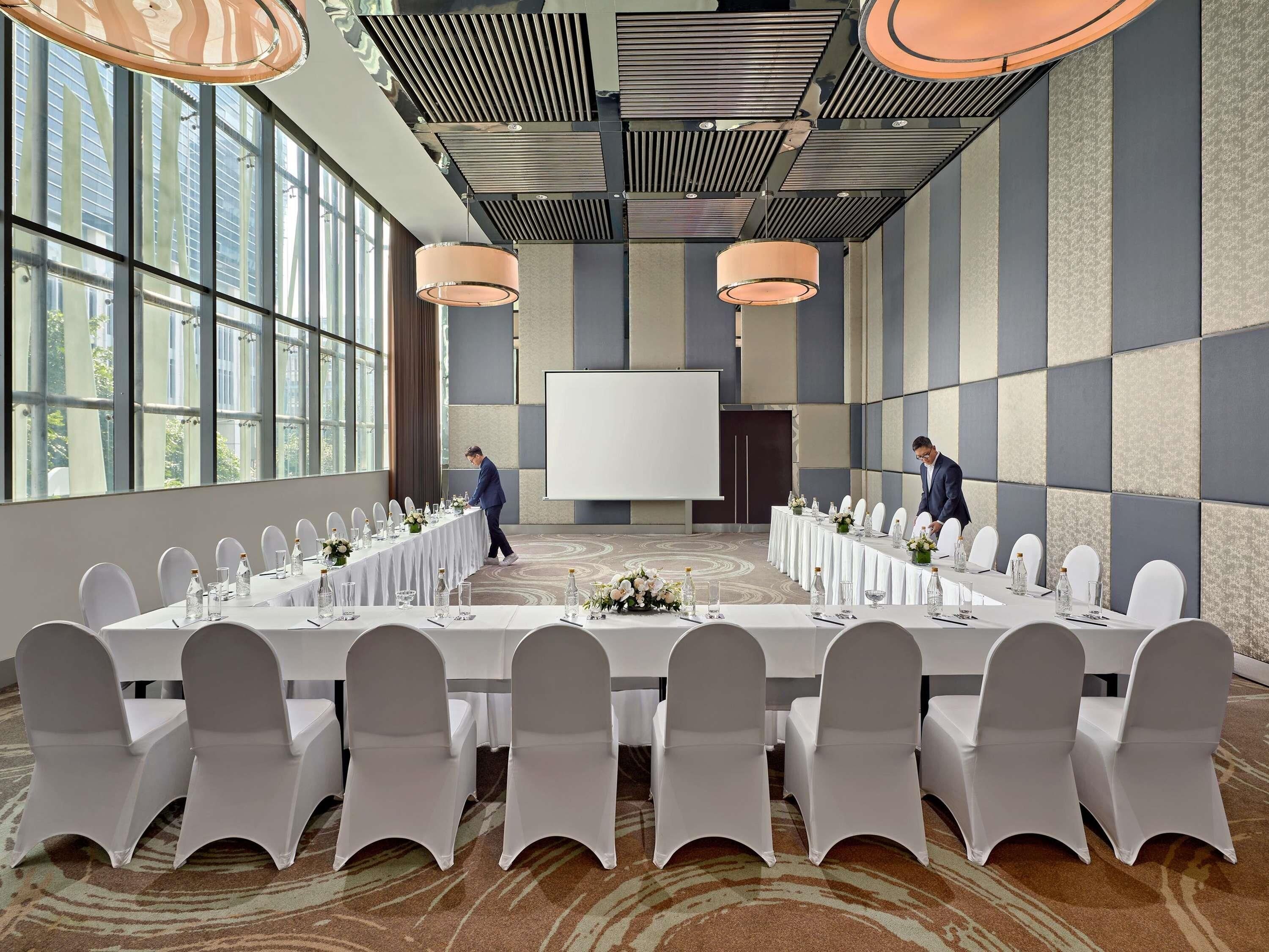 Een vergaderzaal met lange tafels, witte stoelen en een groot scherm.