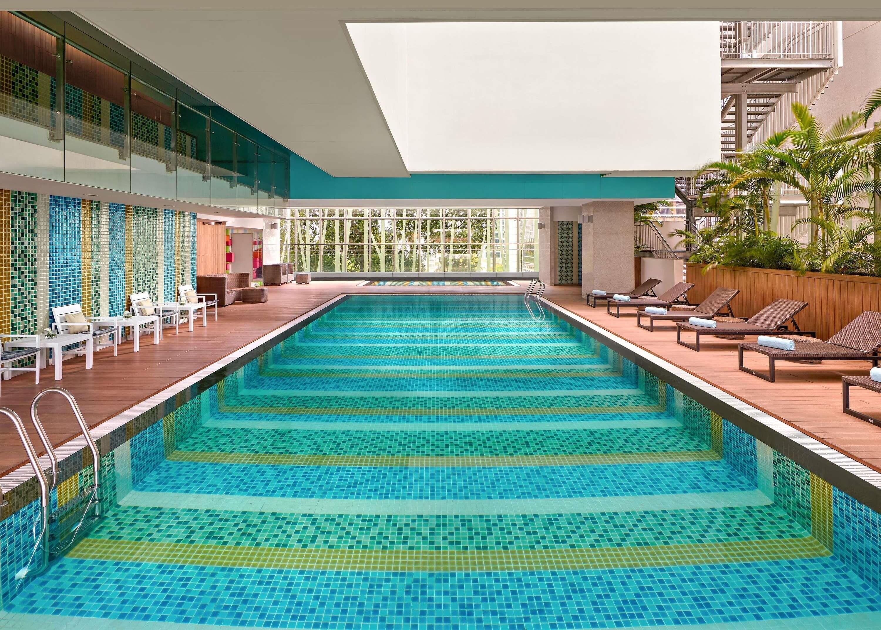 Una piscina interior moderna con agua azul cristalina, sillas de descanso y vistas a un edificio con balcones.