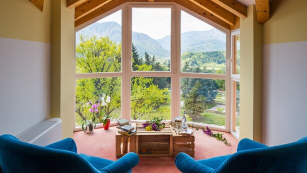 A room with a large window, wooden beams, a radiator, a table with books and flowers, and blue chairs. - Nature Bio Hotel Elite (Levico Terme)