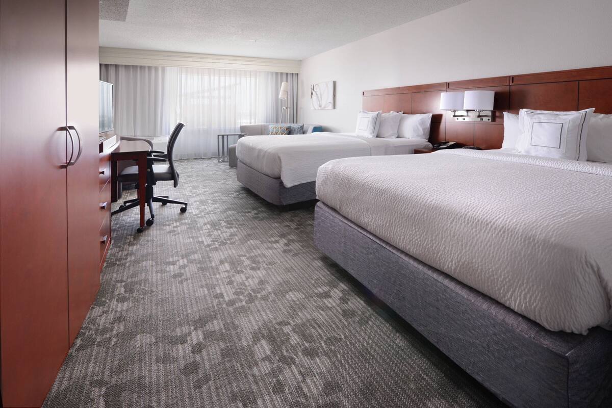 A hotel room with two beds, a desk with a chair, a television, and a large window with curtains.
