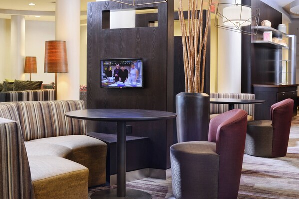 Un moderno vestíbulo de hotel con un televisor montado en una pared oscura, asientos cómodos y un jarrón decorativo con pastos altos.