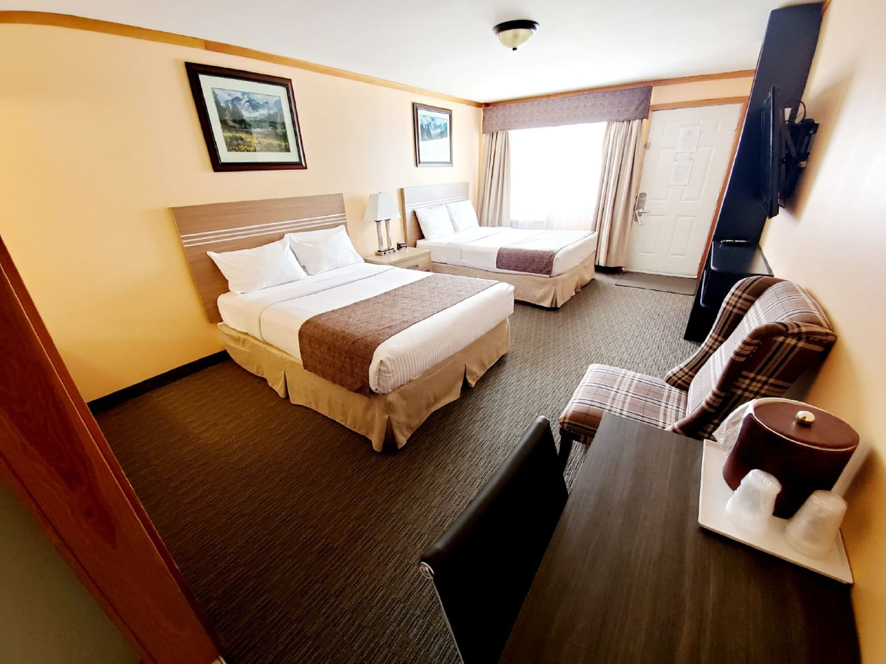A hotel room with two beds, a desk, a chair, a table with a cup and sugar cubes, and a door.