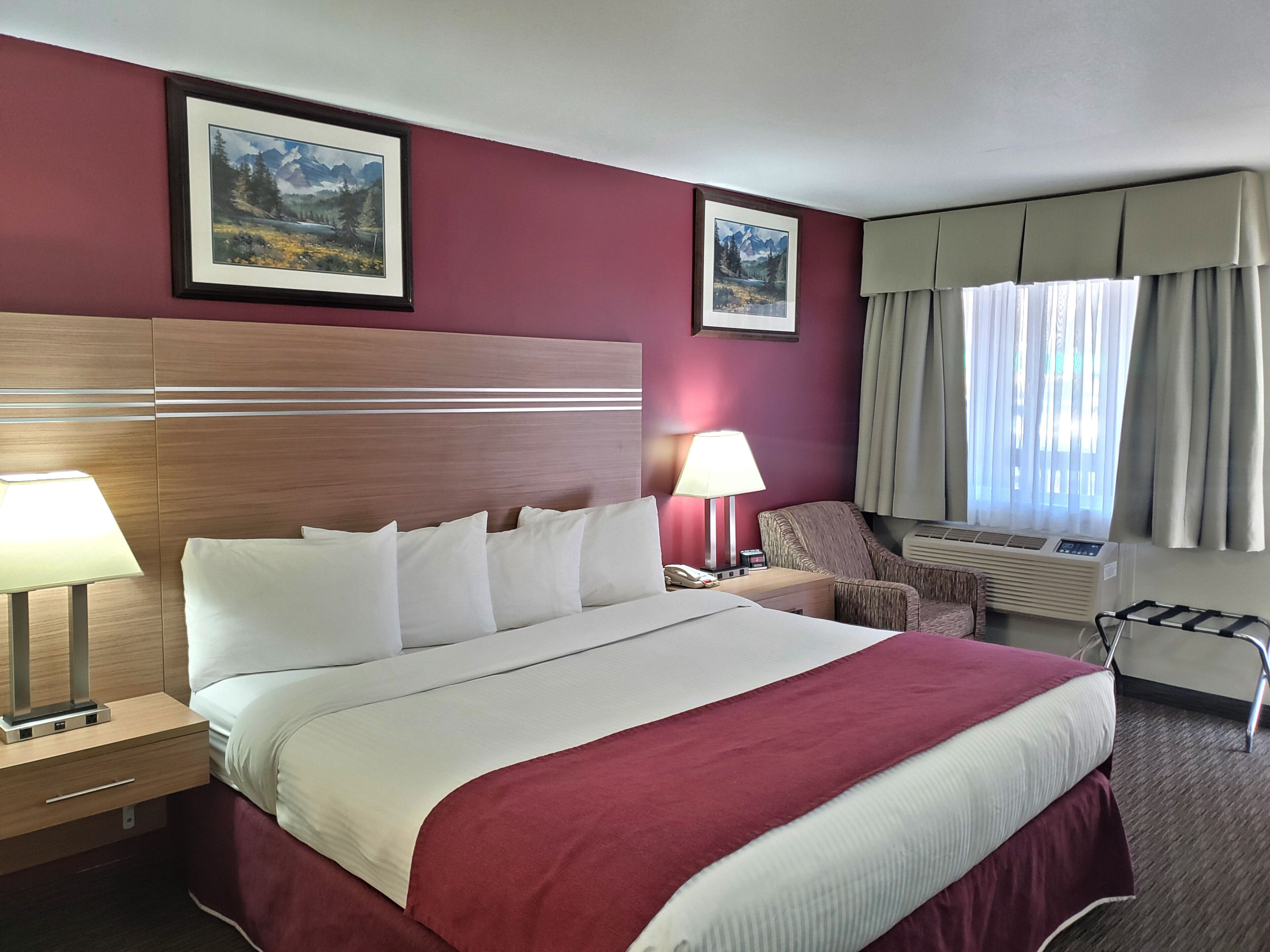 Una habitación de hotel con una cama grande, cabecera de madera, dos cuadros enmarcados en la pared, una silla, un escritorio con lámpara y una ventana con cortinas.
