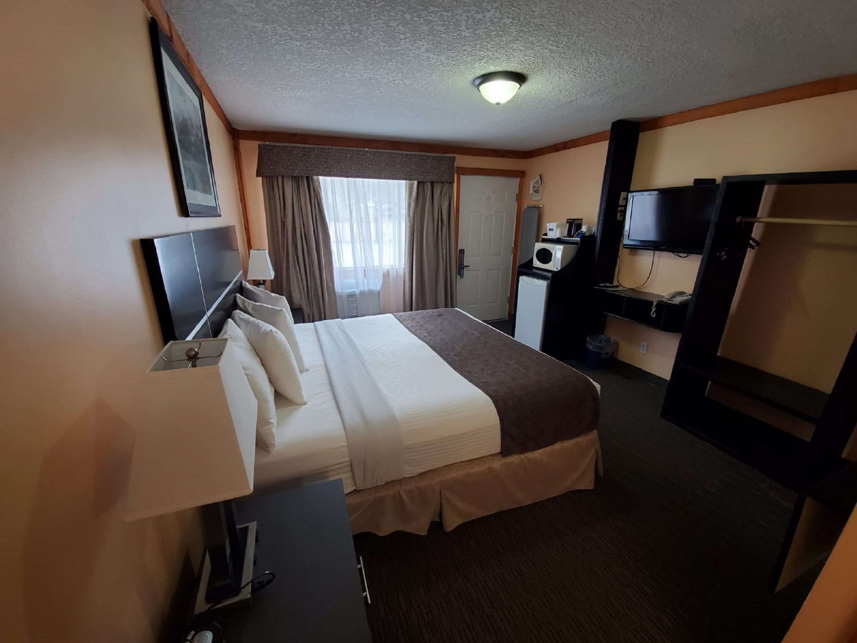 Habitación de hotel con cama, mesita de noche, lámpara, televisor, armario y una pequeña kitchenette.