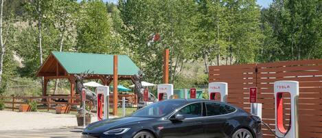 Un coche eléctrico Tesla cargando en una estación con una estructura de madera y árboles al fondo.