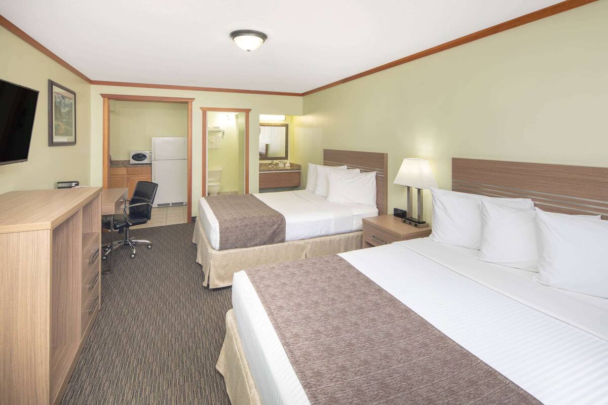 A hotel room with two beds, a desk, a chair, a TV, a bathroom, and a small kitchenette.