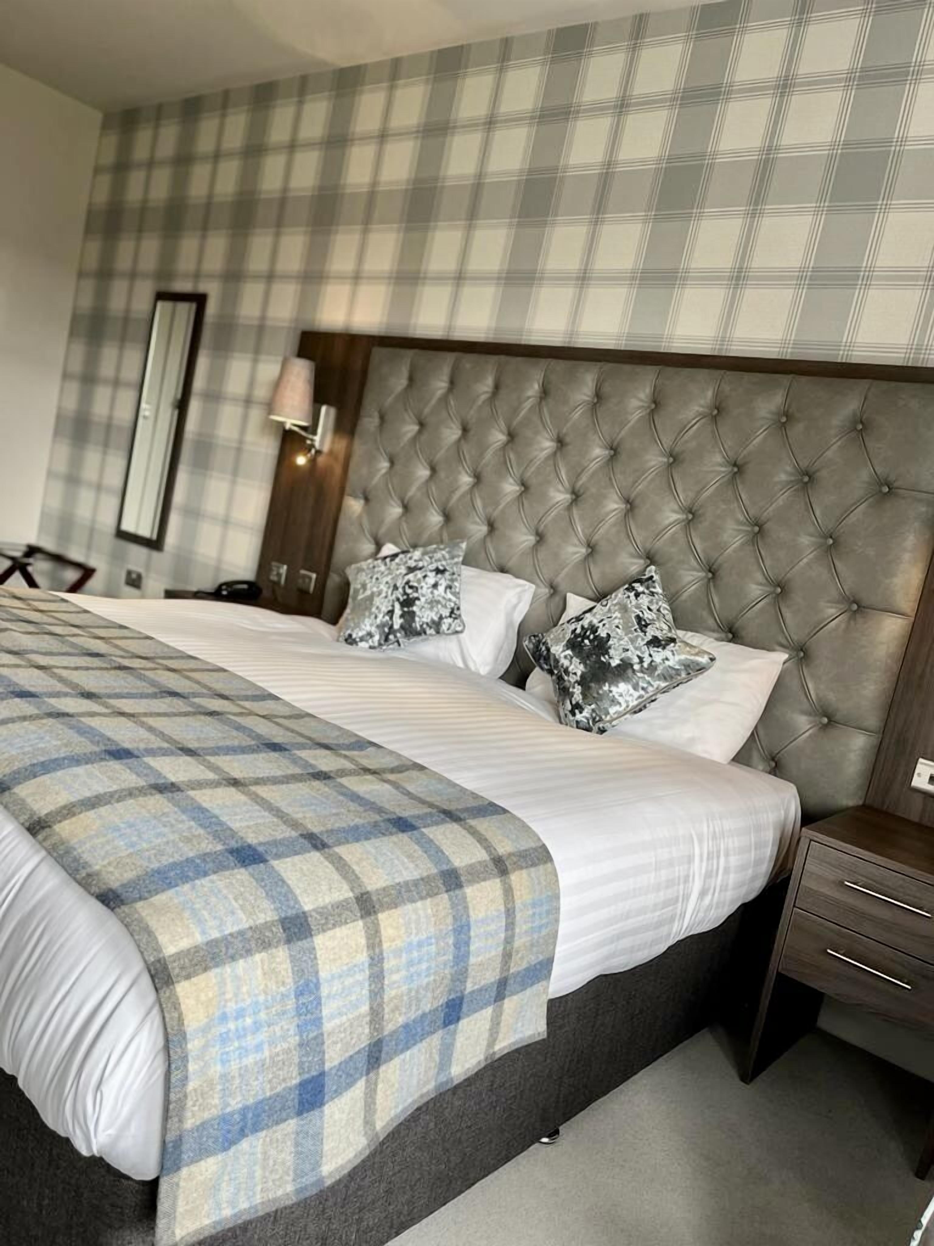 A hotel room with a large bed, a wooden headboard, a nightstand, a lamp, and a plaid bedspread.