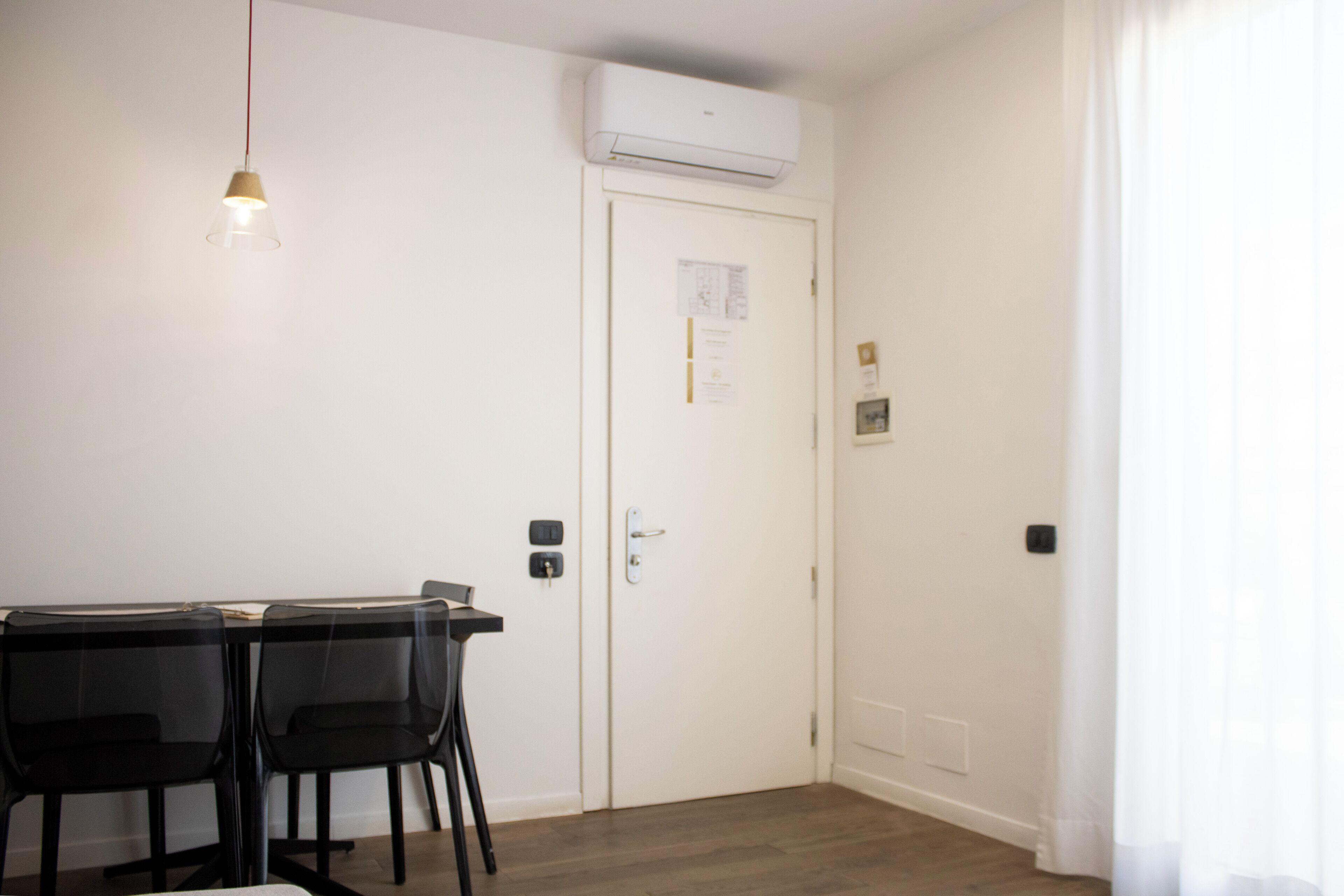A room with a dining table and chairs, a hanging light fixture, a white door with a notice, and an air conditioning unit above.