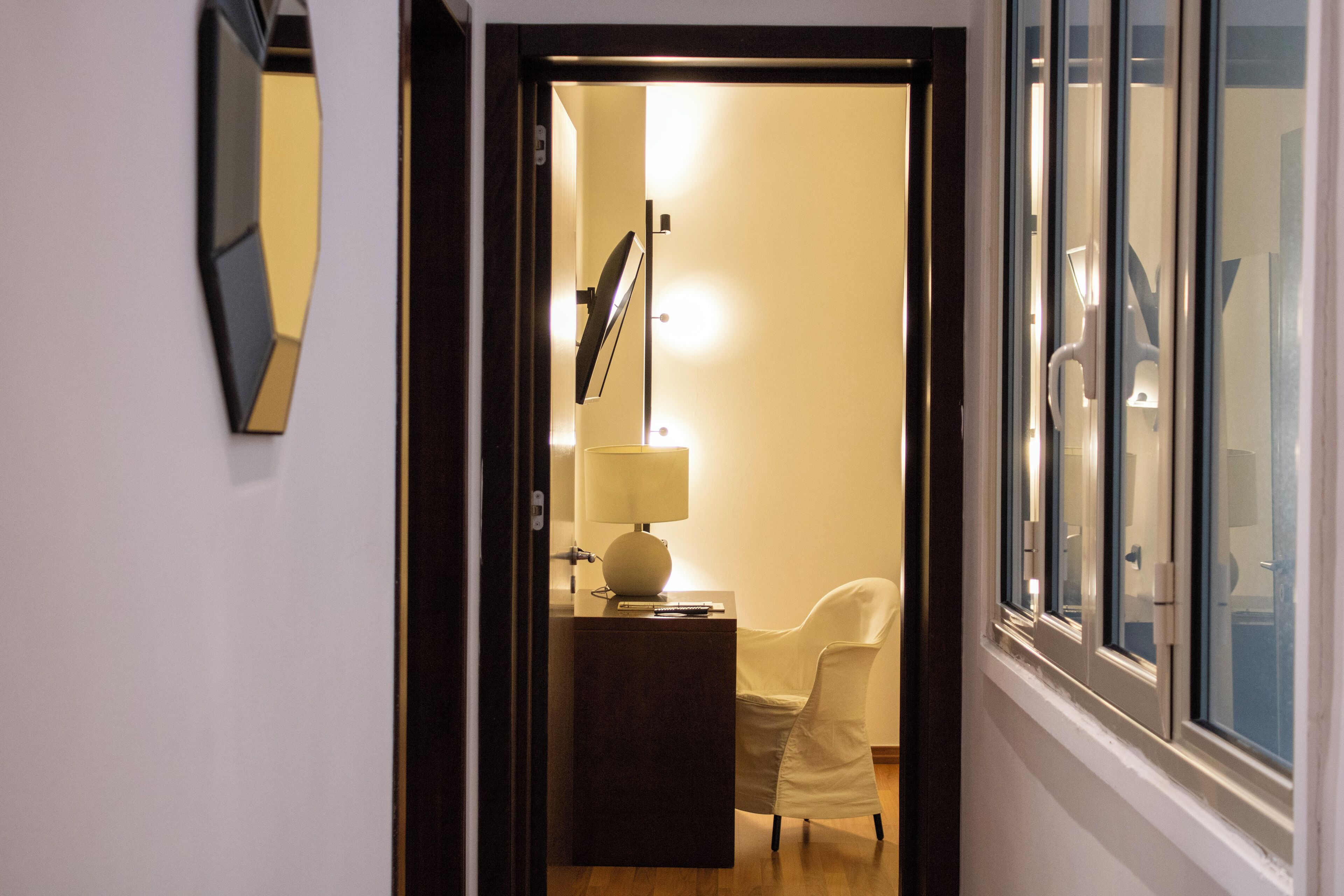 A hallway leading to a room with a desk, chair, lamp, and a large window.